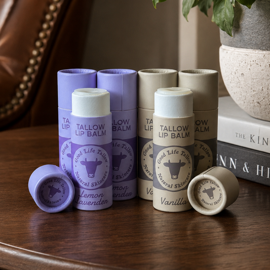 Three lip balm tubes and two loose caps from Tallow Lip Balm are arranged on a wooden table in a styled interior setting with a leather chair, books, and a potted plant in the background.