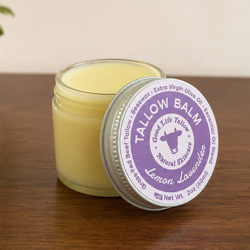 A small glass jar of yellow balm with its silver lid leaning against it on a wooden table. The lid label is white and purple and reads 'TALLOW BALM Good Life Living Natural Lemon Lavender.